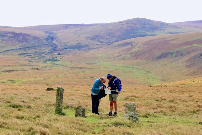 Two Dartmoor walkers study maps next to a standing stone on Dartmoor with a large, moorland Tor in the background.