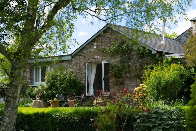 The gable end of a small, stone-built, cottage overlook a garden with a lawn, shrubs and overhanging trees.