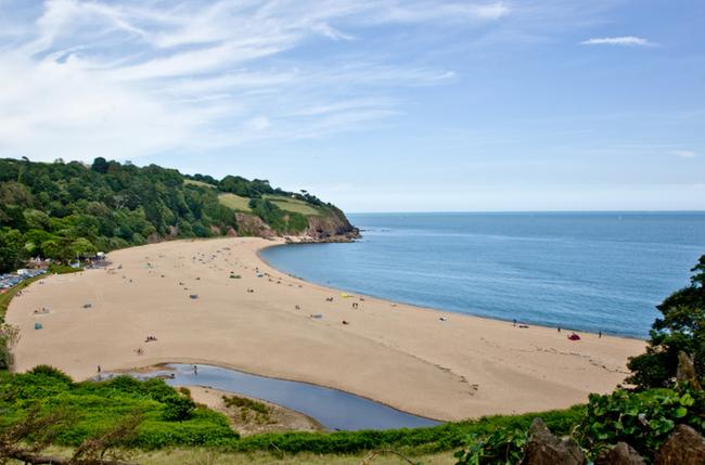 A large sandy cove overlooked by steep-banked wooded hills and cliff tops in South Devon