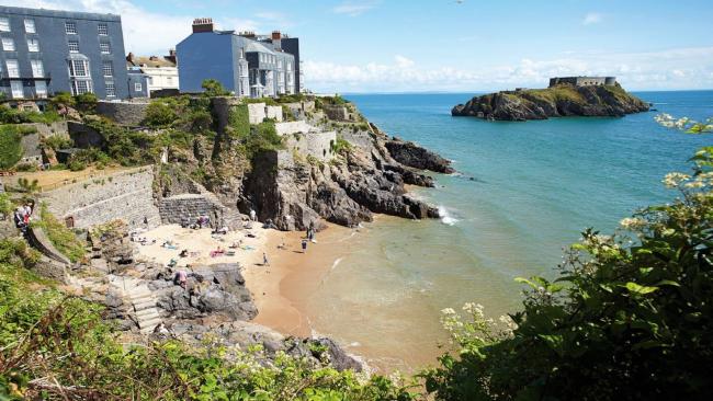 Tall stone houses on a rocky cliff edge overlook a small sandy beach and the ocean.